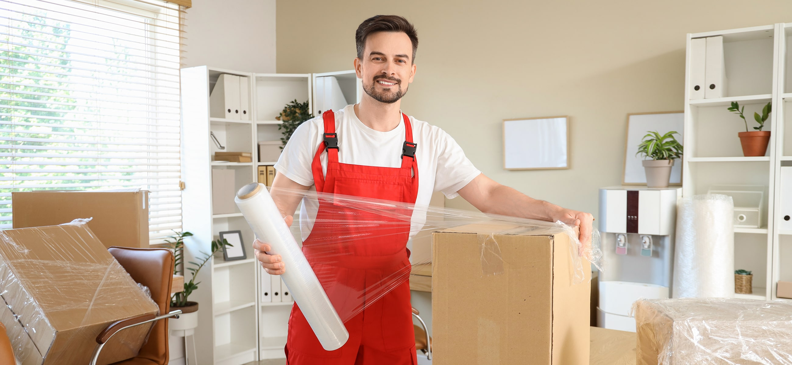 ein Umzugshelfer steht in einem Zimmer und verpackt einen Umzugskarton in Folie
